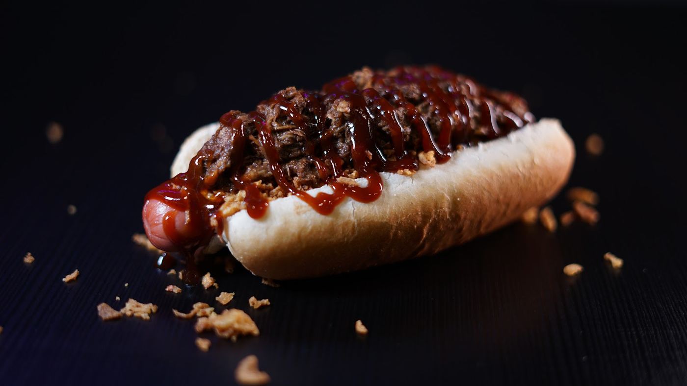 Hot Dog Barbacoa con carne desmechada y salsa casera, servido en pan brioche.